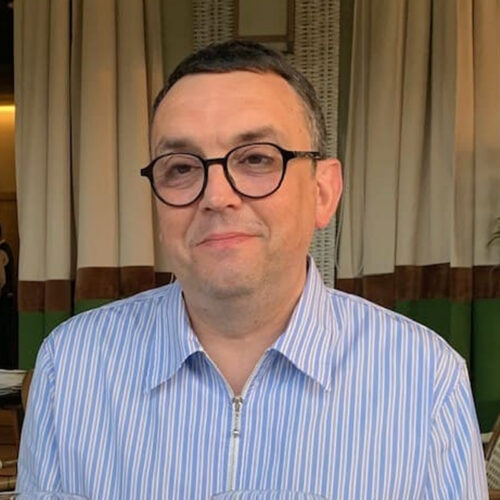 Portrait of Bruno Witek wearing glasses and a striped shirt, seated indoors.