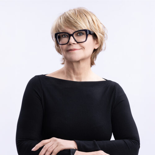 Portrait of Isabelle Witek wearing glasses and a dark sleeveless top, seated in front of a modern office background.