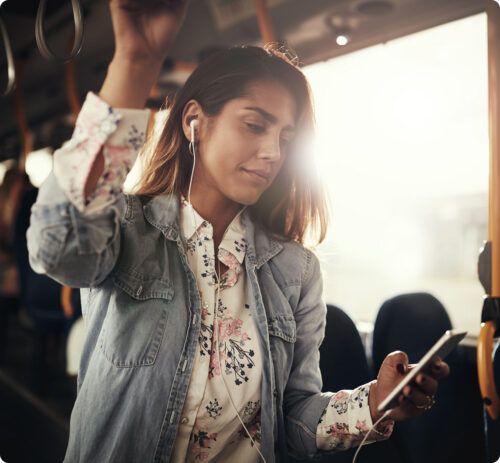 Young woman wearing earphones and holding a smartphone while standing on a bus or train.