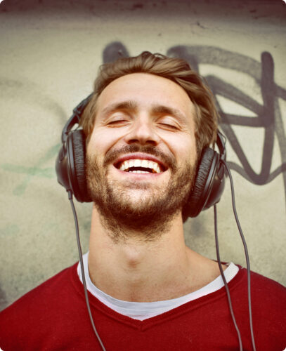 Young man with closed eyes smiling while listening to music through headphones against a graffiti wall.