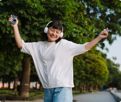 Young woman wearing headphones dancing in a park while holding a smartphone.