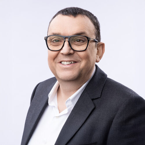 Portrait of Bruno Witek wearing glasses and a striped shirt, seated indoors.